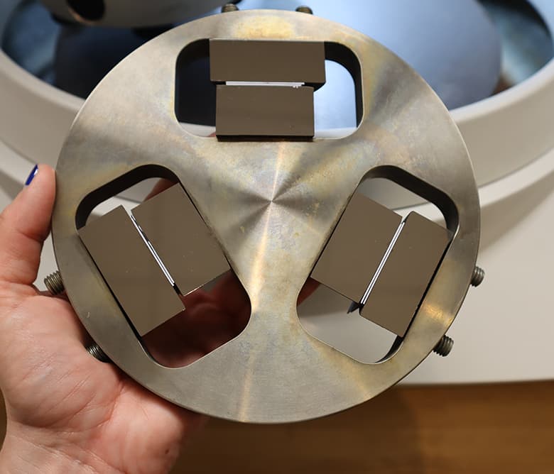 Sample holder designed for metallographic preparation, featuring three evenly spaced sample slots in a circular metal frame. The holder is shown in hand with a polishing machine in the background.